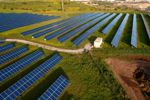 Aerial drone view of solar panels installed on countryside field. Rows of blue photovoltaic panels capture sunlight for renewable energy. Green grass surrounds installation, with dirt roads passing through