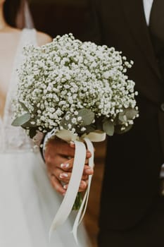 Hands of a Caucasian bride and groom together holding a beautiful wedding bouquet of white boutonnieres on a summer day, close-up side view with selective focus and depth of field.