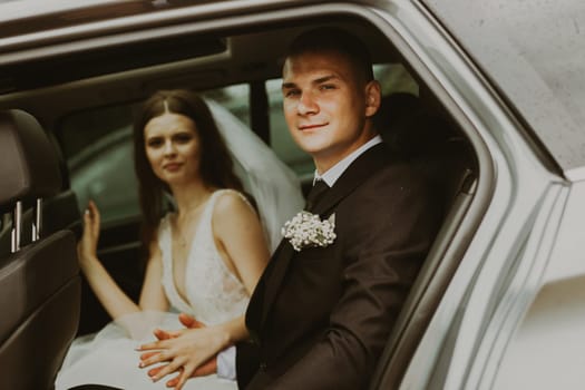Portrait of beautiful young Caucasian happy bride and groom holding each other's hands and sitting inside a car on a summer rainy day looking at the camera, side view close up with selective focus.