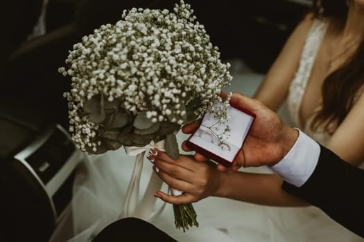 Portrait of beautiful young Caucasian unrecognizable bride and groom who are holding wedding rings box and wedding bouquet of flowers boutonnieres in their hands sitting inside a car on a summer rainy day, close-up side view with selective focus.