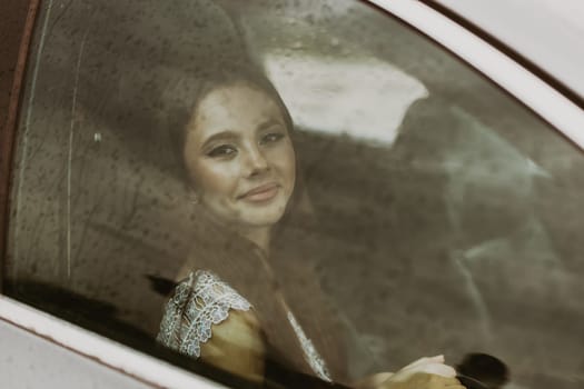 Portrait of one young beautiful Caucasian teenage girl with festive makeup sits inside a car and looks at the camera with a smile on a rainy and cloudy summer day, close-up side view.