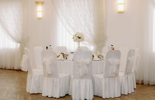Beautiful view of a white round table without people with chairs with painted bows on the backs and a wedding setting of food and drinks on the table in a banquet hall on a summer day, mid shot side view.
