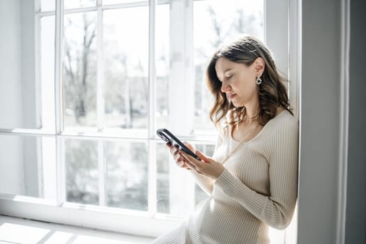 A woman sits against a wall beside a large window, absorbed in her smartphone amidst a bright atmosphere created by natural sunlight.