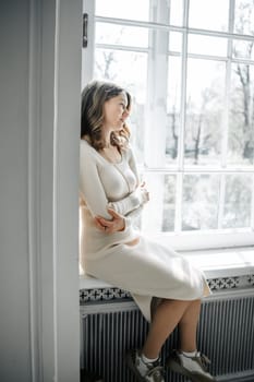A woman in a knitted dress sits comfortably by a large window, enjoying the natural light and serene atmosphere of the indoor space.