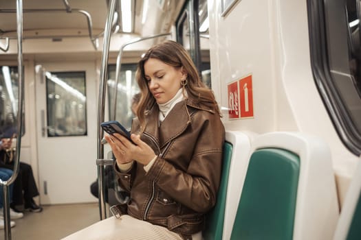 A young woman in a brown leather jacket is focused on her smartphone while sitting alone on a train during rush hour, surrounded by commuters.
