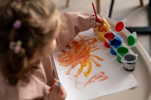 Focused little girl sitting at a kids table, carefully painting with a brush and gouache paints. Indoor creative activity, developing fine motor skills.