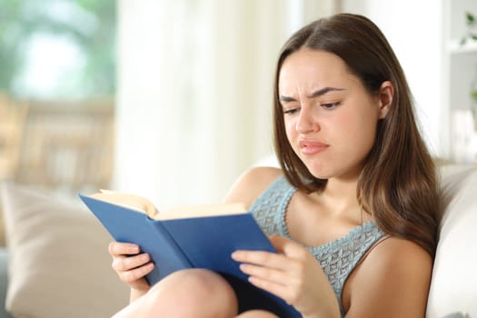 Disappointed woman reads a paper book sitting in a couch at home