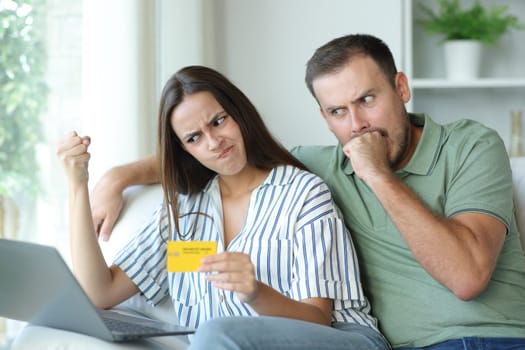 Angry couple paying online with laptop and credit card sitting in a couch at home