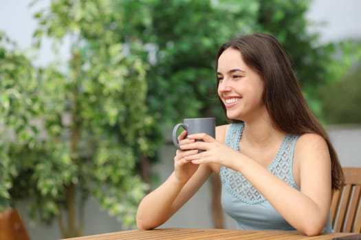 Happy woman holding coffee cup looking away sitting in a terrace