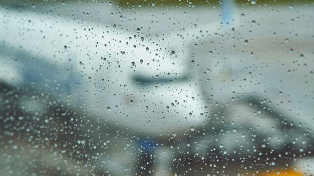 Raindrops clinging to a window obscure the view of an airplane on the tarmac, creating a moody atmosphere at the airport