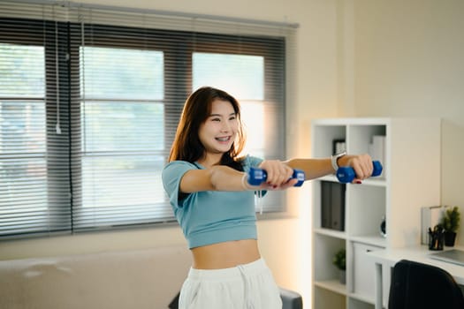 Cheerful young woman exercises with small dumbbells in her living room.