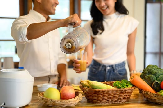Smiling couple pouring fresh blended smoothie into a glass in a cozy kitchen.