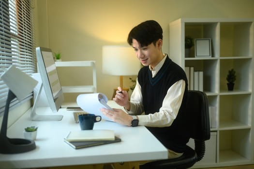 Young man reviewing documents at home office with computer and coffee nearby.