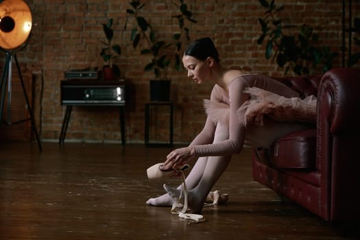 A poised ballerina is gently and delicately wearing her ballet shoes inside a beautifully stylish studio ambiance