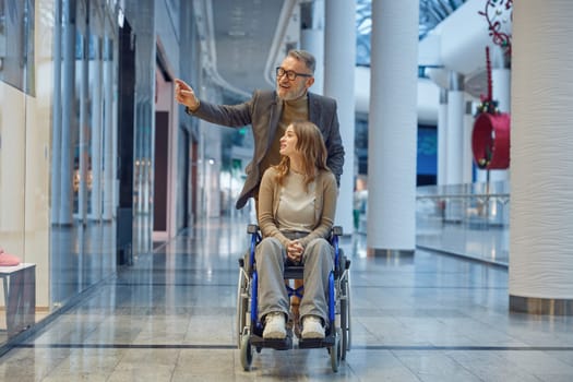Man gently pushes woman in wheelchair along long, spacious hallway that seems to stretch before them