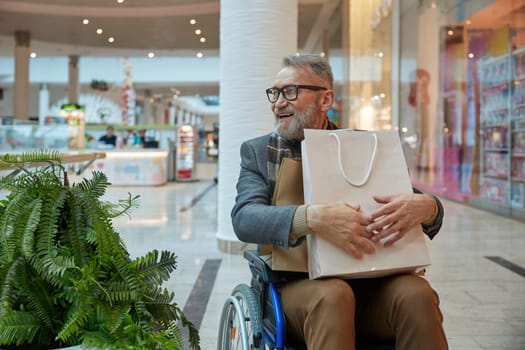 A man who is seated in a wheelchair holding shopping bag while moving through bustling mall filled