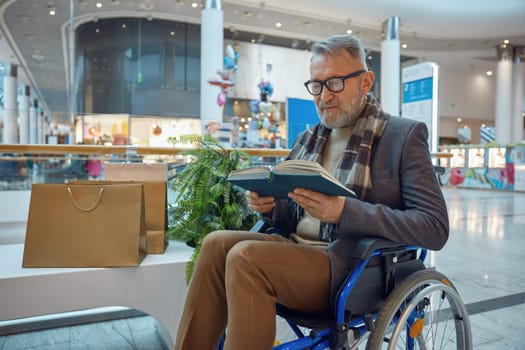 Man who uses wheelchair comfortably seated on bench and is engaged in reading an interesting book as he enjoys his day