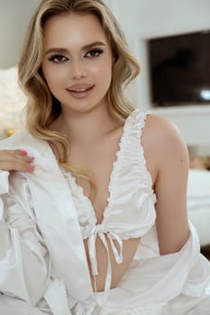 portrait of beautiful young blonde with long, wavy hair in silk white robe and white underwear. she looks at camera and smiles. close-up of well-groomed girl. girl sits on white bed in light interior