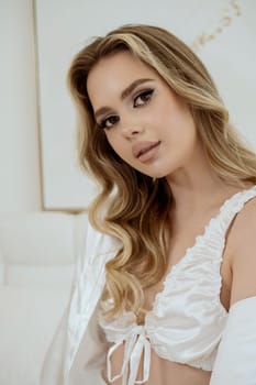 portrait of a beautiful young blonde with long, wavy hair in a silk white robe and white underwear. close-up of a well-groomed girl, she looks at the camera