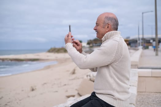 An elderly person sitting by the ocean shore, taking photos of the view. Active retirement lifestyle.
