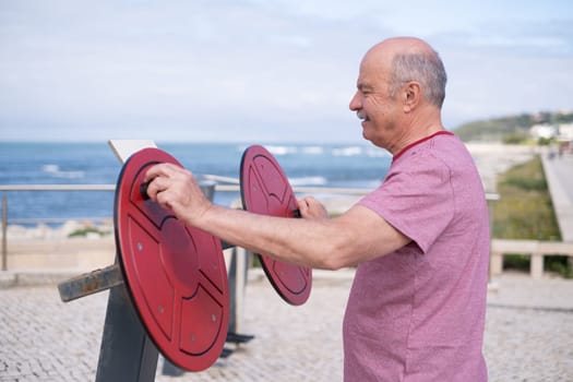 An elderly man stretching his arms on an outdoor fitness machine by the ocean shore. High quality photo