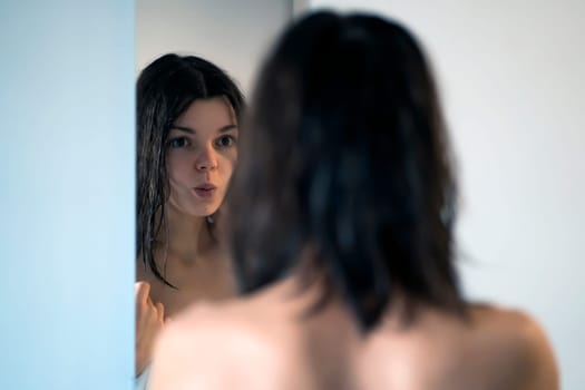 After a shower and hygienic procedures, a young pretty girl, wrapped in a towel, looks at herself in the mirror, applies a mask on her hair, does skin care at home.