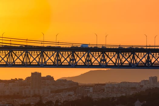 Beautiful view of golden sunset and suspension city bridge with its steel structure and cables, dramatic silhouettes of vehicles in motion blur against bright orange sky. Distant hills and urban skyline of modern buildings in evening summer haze.