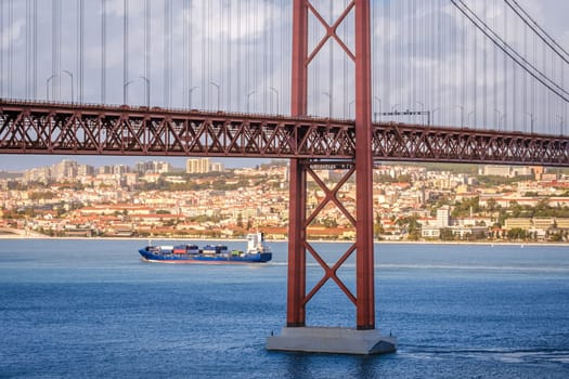 Colorful landscape with blue container ship on Tagus river under iconic 25 de Abril or 25th April bridge, Lisbon, Portugal. Famous ironwork of red suspension bridge, vibrant cityscape, urban architecture, maritime transport, sunny day, cloudy sky