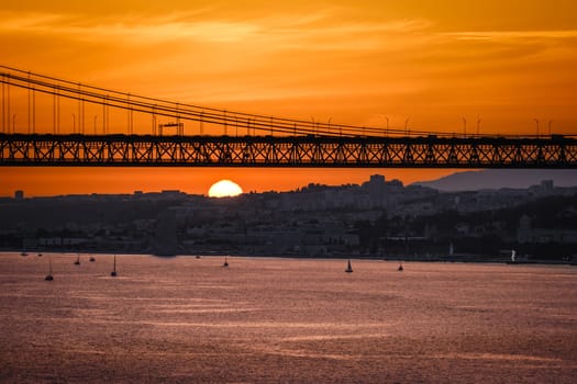 Beautiful sunset over 25 de Abril bridge in silhouette and Tagus river with sailboats drifting below and Lisbon skyline in summer haze in the background. Orange sky, clouds and sun, tranquil waters, traffic on bridge, iconic view of Lisbon, Portugal.