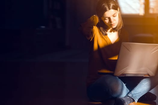 The girl sits on the floor of a cozy house in the rays of the sun and chats, works on a laptop online. A woman learns and develops her business at home.