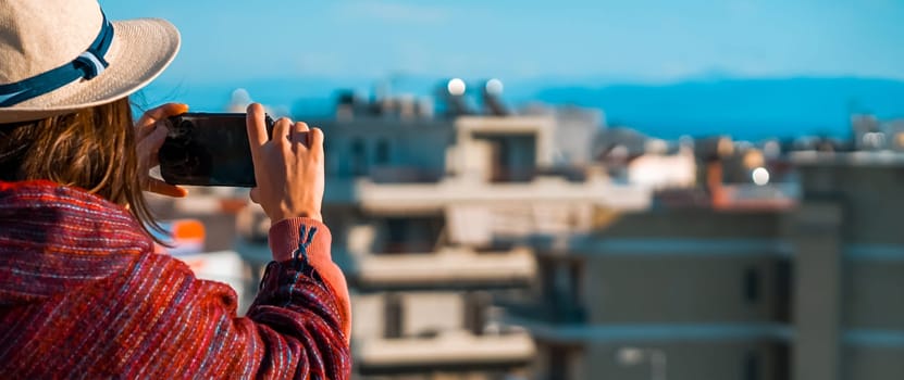 A young girl in a burgundy poncho and a hat travels and takes a photo with a cityscape on a sunny day.