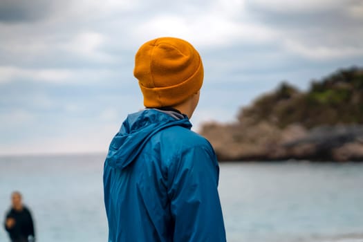 A man in a windbreaker and a yellow hat looks towards the ocean in atmospheric cool weather, a tourist backpacker travels along the northern coast along the sea...