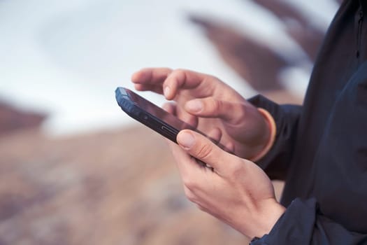 Man holds a smartphone in his hands and looks at a map, makes a route through the mountains, writes a message, chats. Young traveler explores the area, uses the app on his device while hiking.