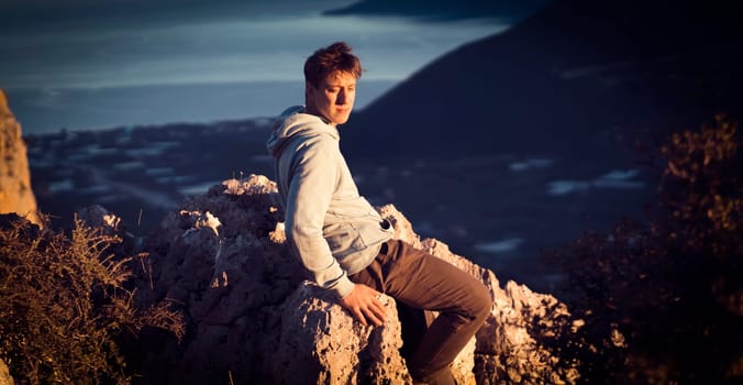 A man sits on a stone on top of a mountain in the sun, against the backdrop of a beautiful landscape with a sea coast and a small cozy city, the traveler is trekking during his trip.