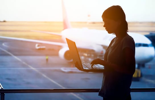A young girl works on a laptop at the airport while she waits to board a plane. A woman buys tickets, studies and communicates via the Internet, the airport in the background.