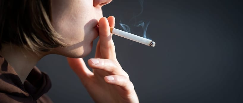 A young beauty girl is smoking, a woman is stressed and holds a cigarette in her hands, staying near the window of her apartment, close-up view.