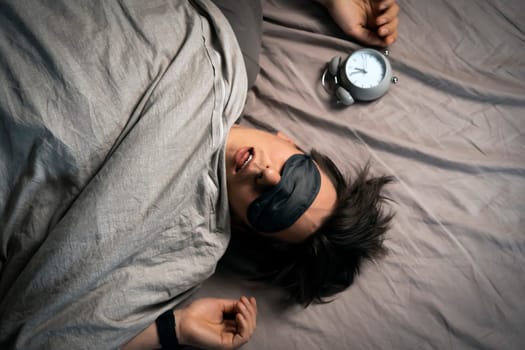 A young funny man sleeps soundly early in the morning, hugs a pillow and holds a vintage alarm clock in his hands, the sleep mask has slipped from his eyes.