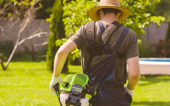 A young man in a straw hat is mowing a lawn with a lawn mower in his beautiful green floral summer garden. A professional gardener with a lawnmower cares for the grass in the backyard.