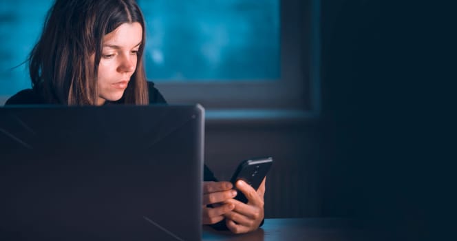 A young girl works at home at night with a laptop, a woman holds a phone in her hands, participates in a conference meeting online, studies, searches for information using her device.