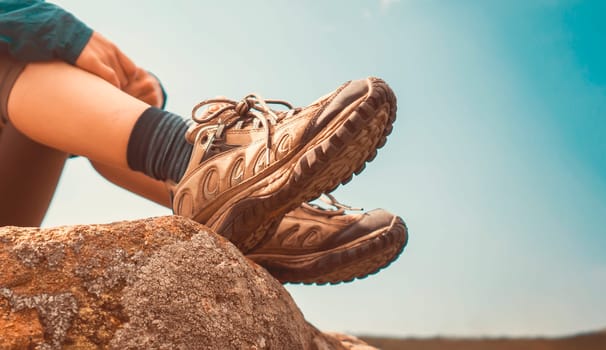 A beautiful girl in a hat sits on a rock against the sky, legs in trekking sneakers close-up. A young woman travels, leads an active lifestyle, goes hiking in the mountains.