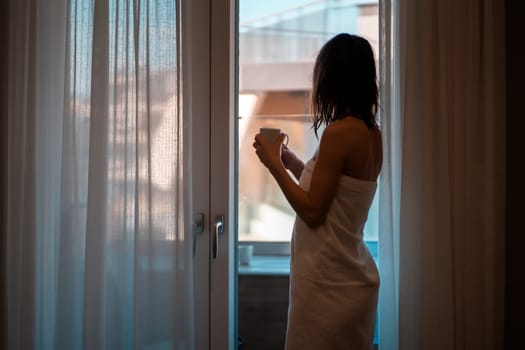 A girl looks out the window after a shower wrapped in a white towel with a cup of coffee, a silhouette of a woman in front of a balcony and curtains on the sides in her cozy room at home.