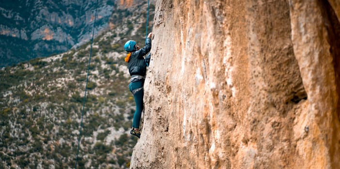 Young girl is engaged in extreme sports, mountaineering, leads an active lifestyle, climbs a rock with climbing equipment, a rope and in a helmet, against the background of hills with olive trees.