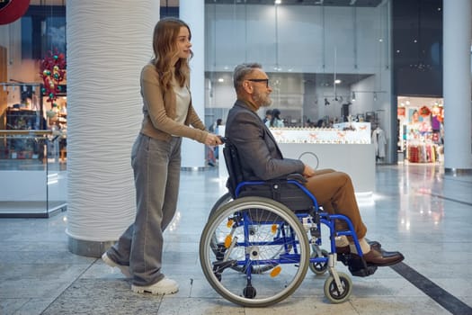 Woman gently pushes a man in wheelchair along a long hallway, filled with soft light