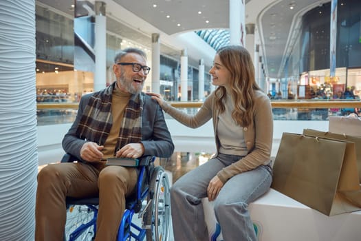 A woman is sitting next to a man who is in a wheelchair in a bustling shopping mall full of various stores and shoppers