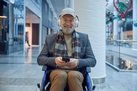 Elderly man in a wheelchair enjoys music while using his smartphone in a contemporary shopping center.