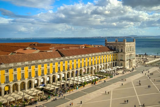 Wide shot of Commerce Square or Praca do Comercio in Lisbon, Portugal, iconic yellow Pombaline architecture, bustling outdoor cafes, Tagus River, city life, historic downtown, sunny day, sky with clouds. Summer vacation, travel destination