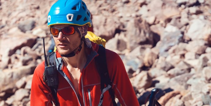 A young man in sunglasses, helmet and a red thermal jacket, with a walkie-talkie walks in the mountains alone. The traveler climbs the rocks, stones, goes in for mountaineering.