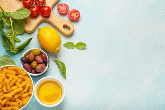 Cherry tomatoes, basil, olives, lemon, olive oil, fusilli pasta and wooden cutting board are creating a frame with copy space on a light blue background, concept of Italian cuisine cooking, copy space