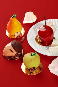 Four mousse cakes in the shape of a pear, apple, cherry, and acorn are beautifully arranged on a red background, creating a visually appealing dessert scene
