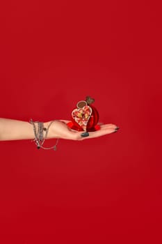 Woman hand with bracelets holding a red heart shaped chocolate praline with a small chocolate heart on top, on a red background
